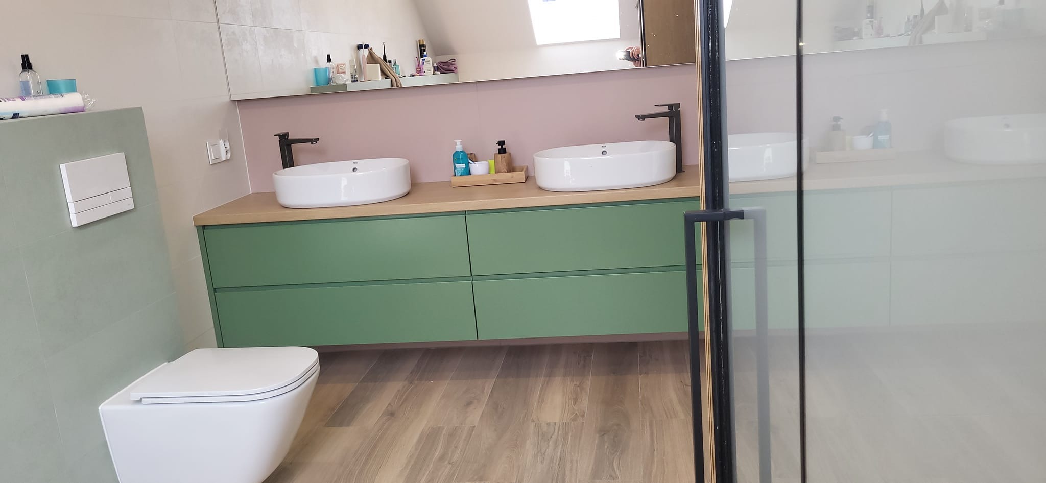 Nowoczesna łazienka z zielonymi szafkami pod umywalkami, drewnianym blatem, białymi umywalkami nablatowymi i wiszącą toaletą.