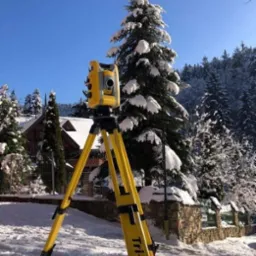 Żółty teodolit Tri-Max ustawiony na śniegu, w tle ośnieżone drzewa i budynek z drewnianą elewacją.