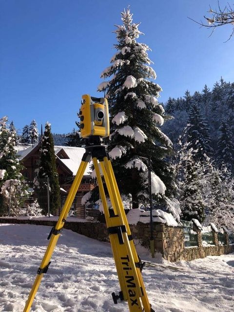 Żółty teodolit Tri-Max ustawiony na śniegu, w tle ośnieżone drzewa i budynek z drewnianą elewacją.