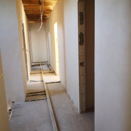 Usługi budowlane Wojciech Szyniec - Długi korytarz w trakcie wykańczania, z gładkimi białymi ścianami, widocznymi drewnianymi belkami stropowymi i instalacjami na betonowej posadzce.