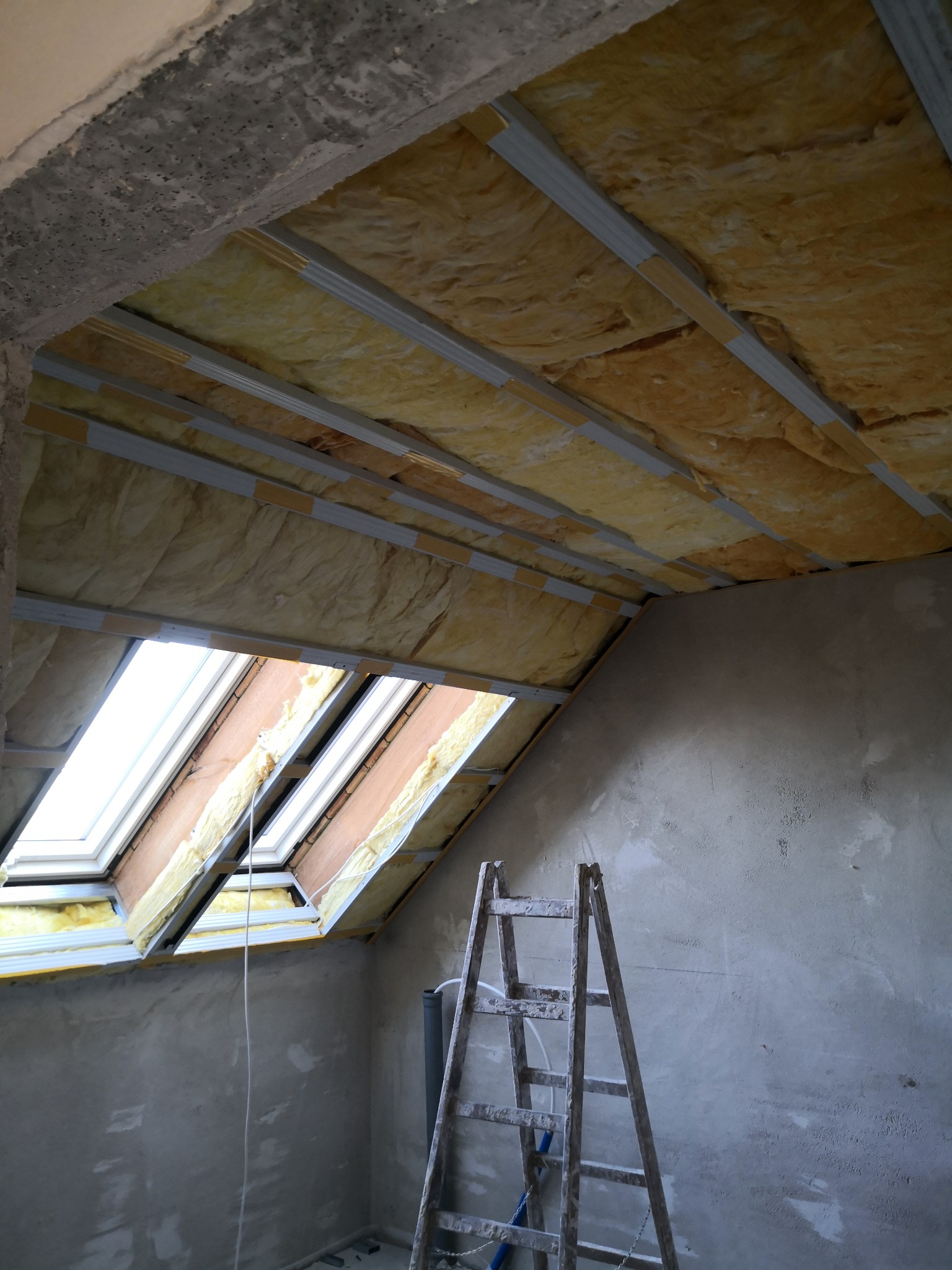 Poddasze w trakcie ocieplania wełną mineralną, widoczne metalowe profile, okno dachowe i drabina oparta o surową ścianę.