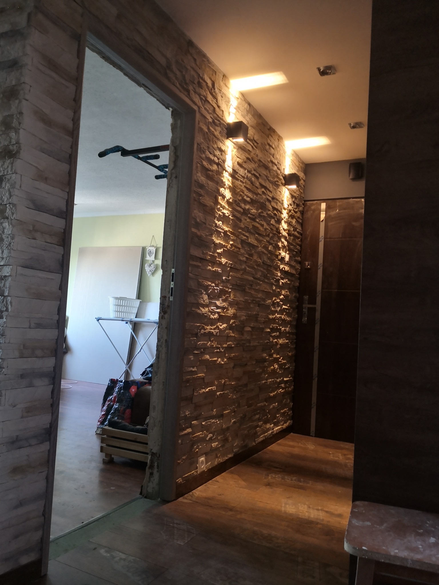Korytarz z dekoracyjną ścianą z kamienia, oświetloną przez czarne, kwadratowe lampy ścienne, widok na pokój z drążkiem do ćwiczeń i deską do prasowania.