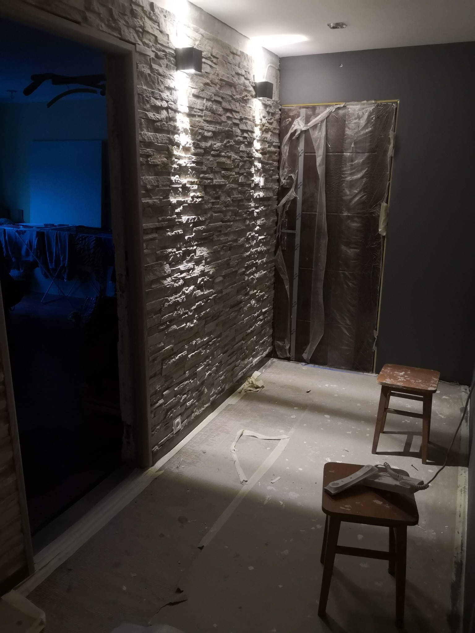 Wykończony fragment korytarza z dekoracyjną ścianą z kamienia, podświetloną kinkietami, z zabezpieczonymi folią drzwiami i dwoma stołkami, na jednym z nich leży telefon stacjonarny.