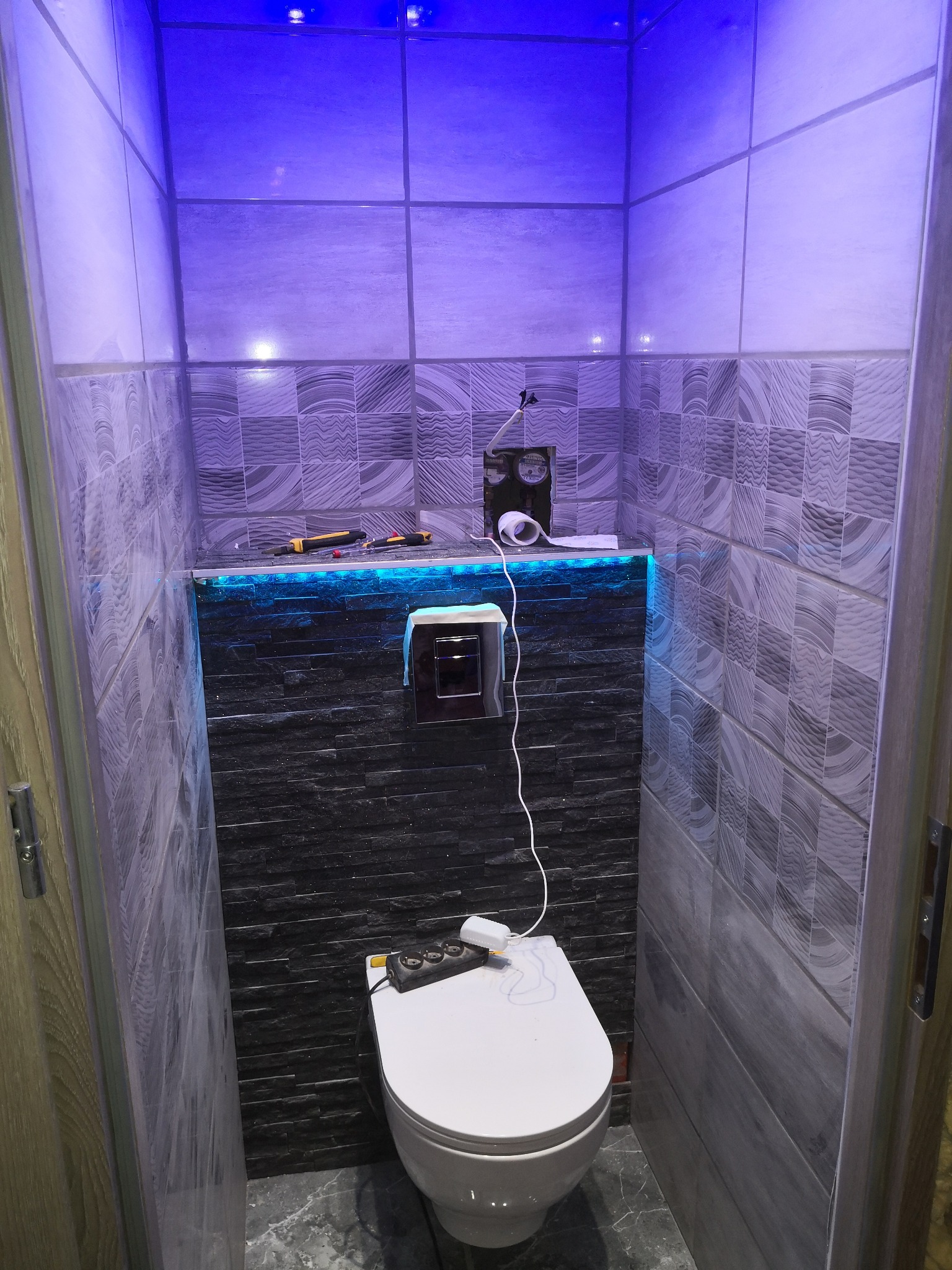 Wnętrze małej łazienki po remoncie z podświetleniem LED, widoczna toaleta, płytki na ścianach w odcieniach szarości i czerni, narzędzia na półce, niebieskie światło sufitowe.