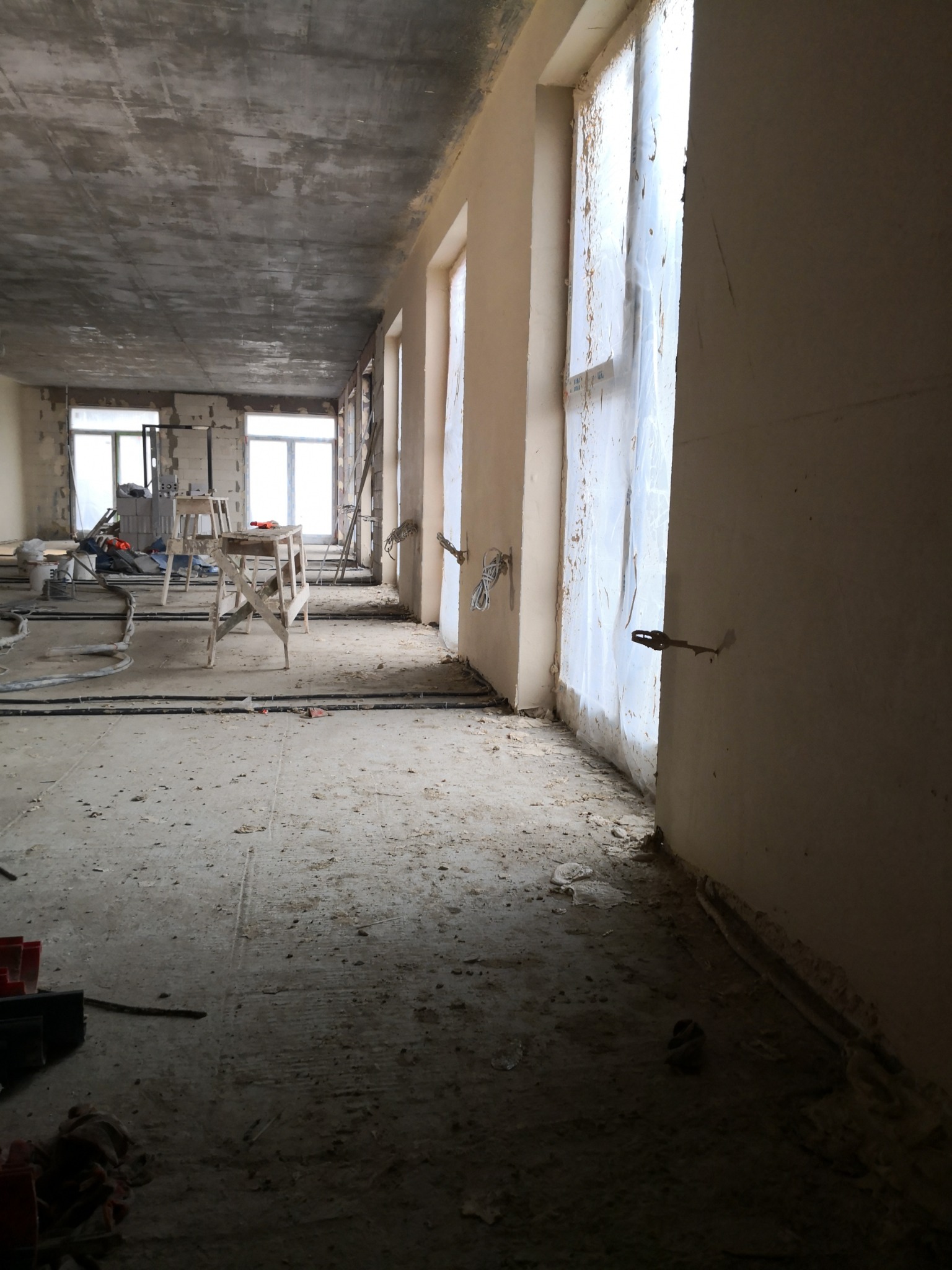 Wnętrze w trakcie remontu: surowy betonowy sufit, ściany przygotowane do wykończenia, okna zabezpieczone folią, widoczne przewody i drabiny malarskie.