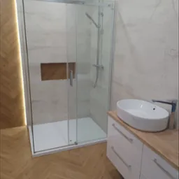 Nowoczesna łazienka z kabiną prysznicową typu walk-in, umywalką nablatową na szafce z drewnianym blatem i podświetlaną wnęką w ścianie. Parkiet w jodełkę na podłodze.