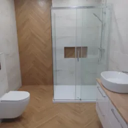 Nowoczesna łazienka z kabiną prysznicową, umywalką nablatową i toaletą. Na ścianie i podłodze drewnopodobna glazura ułożona w jodełkę.