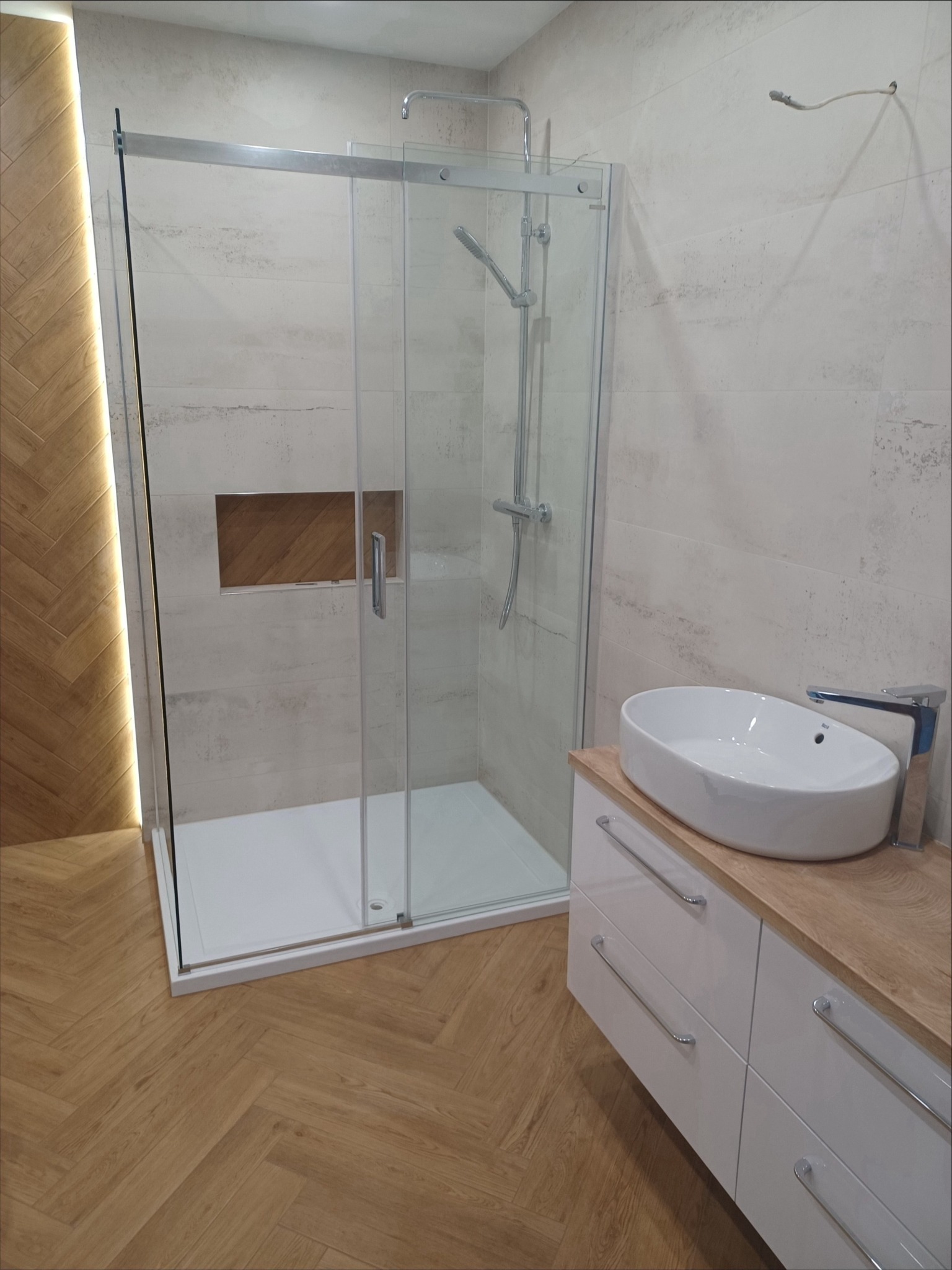 Nowoczesna łazienka z kabiną prysznicową typu walk-in, umywalką nablatową na szafce z drewnianym blatem i podświetlaną wnęką w ścianie. Parkiet w jodełkę na podłodze.