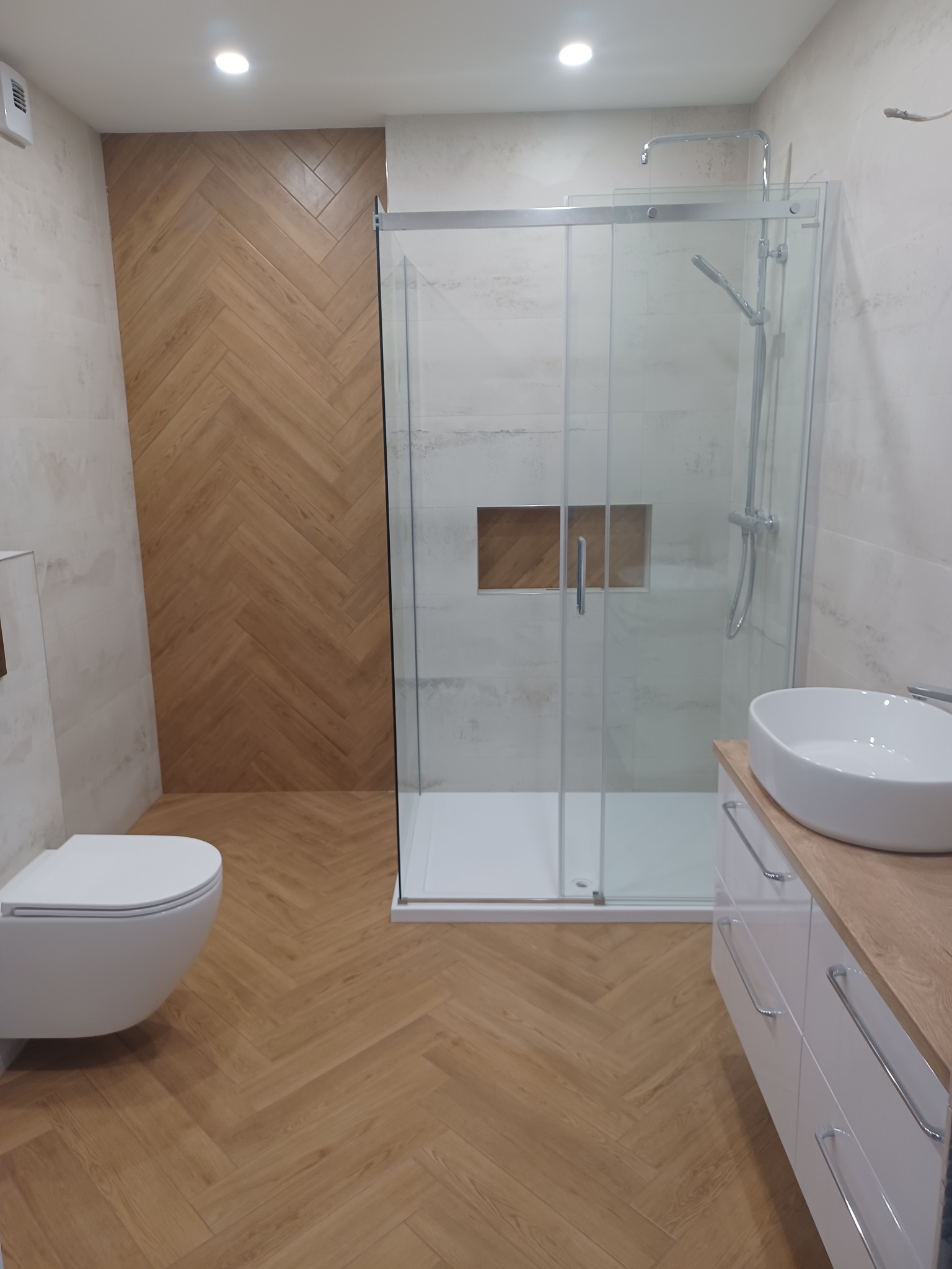 Nowoczesna łazienka z kabiną prysznicową, umywalką nablatową i toaletą. Na ścianie i podłodze drewnopodobna glazura ułożona w jodełkę.