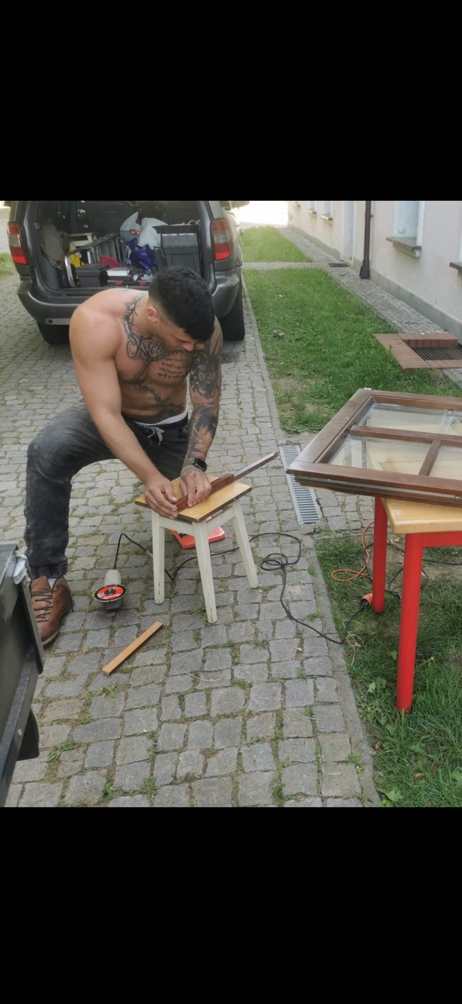 Mężczyzna z tatuażami szlifuje drewniany element ramy okiennej na zewnątrz, z samochodem i narzędziami w tle.