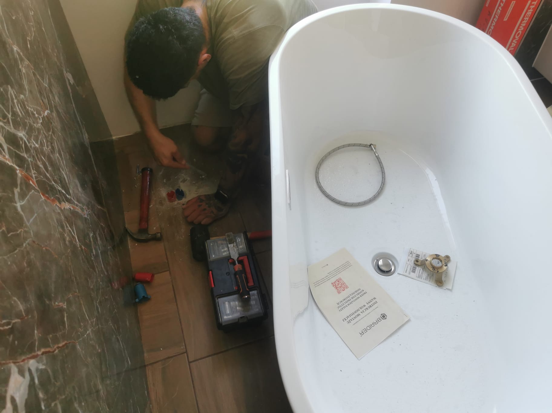 Instalacja wanny wolnostojącej: hydraulik przy pracy, widoczne narzędzia, złączki, instrukcja montażu producenta Braider w wannie, płytki imitujące marmur na ścianie.
