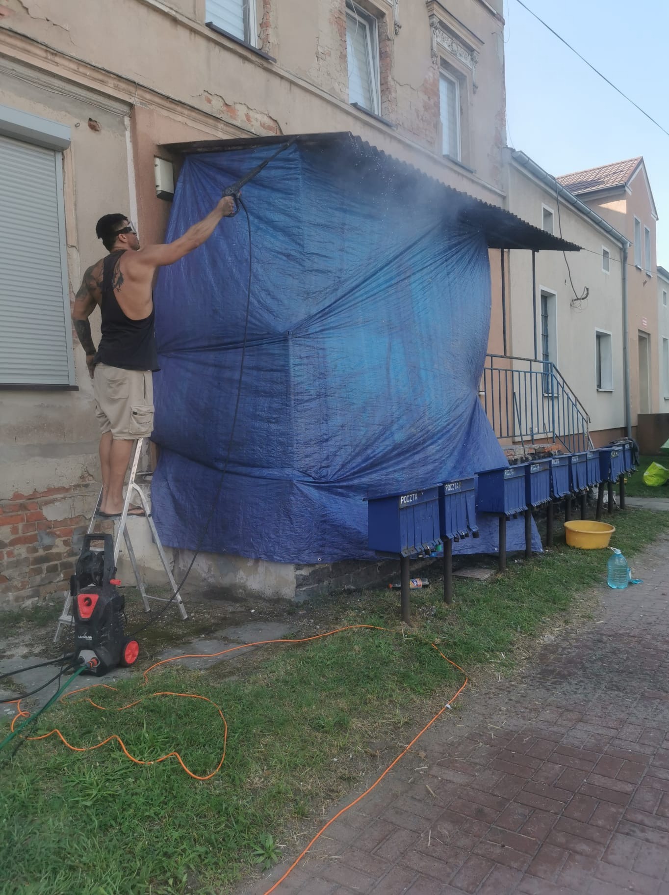 Mężczyzna z tatuażami na drabinie czyści myjką ciśnieniową zadaszenie nad wejściem do budynku, osłonięte niebieską plandeką. Widoczne skrzynki pocztowe i budynek w tle.