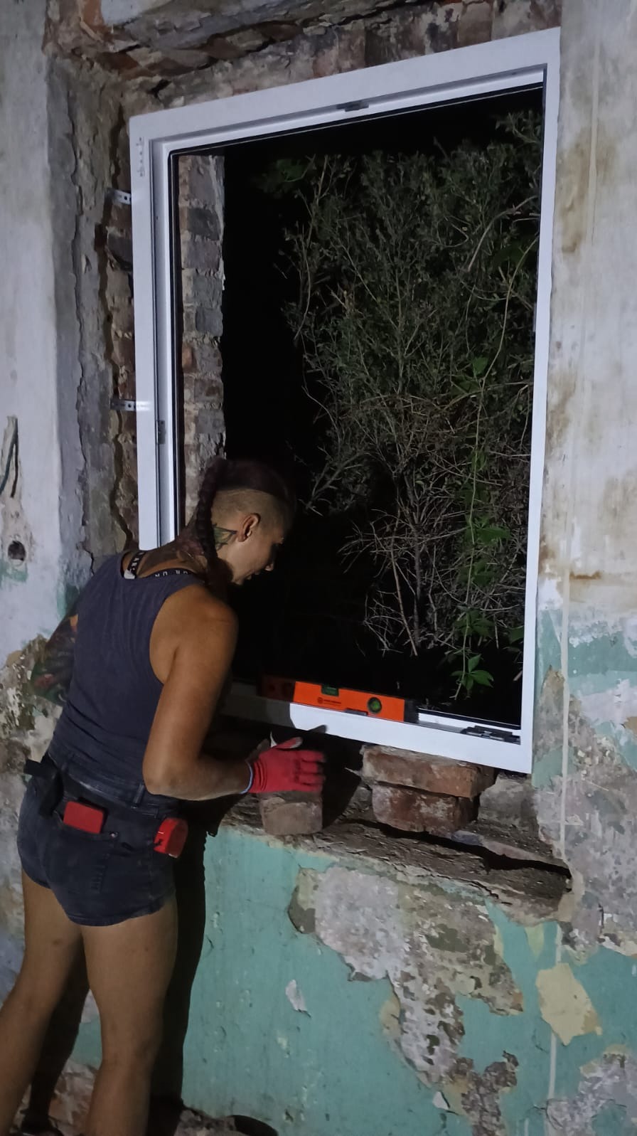 Kobieta w rękawicach montuje białe okno w surowym murze, używając poziomicy i podkładek z cegieł.