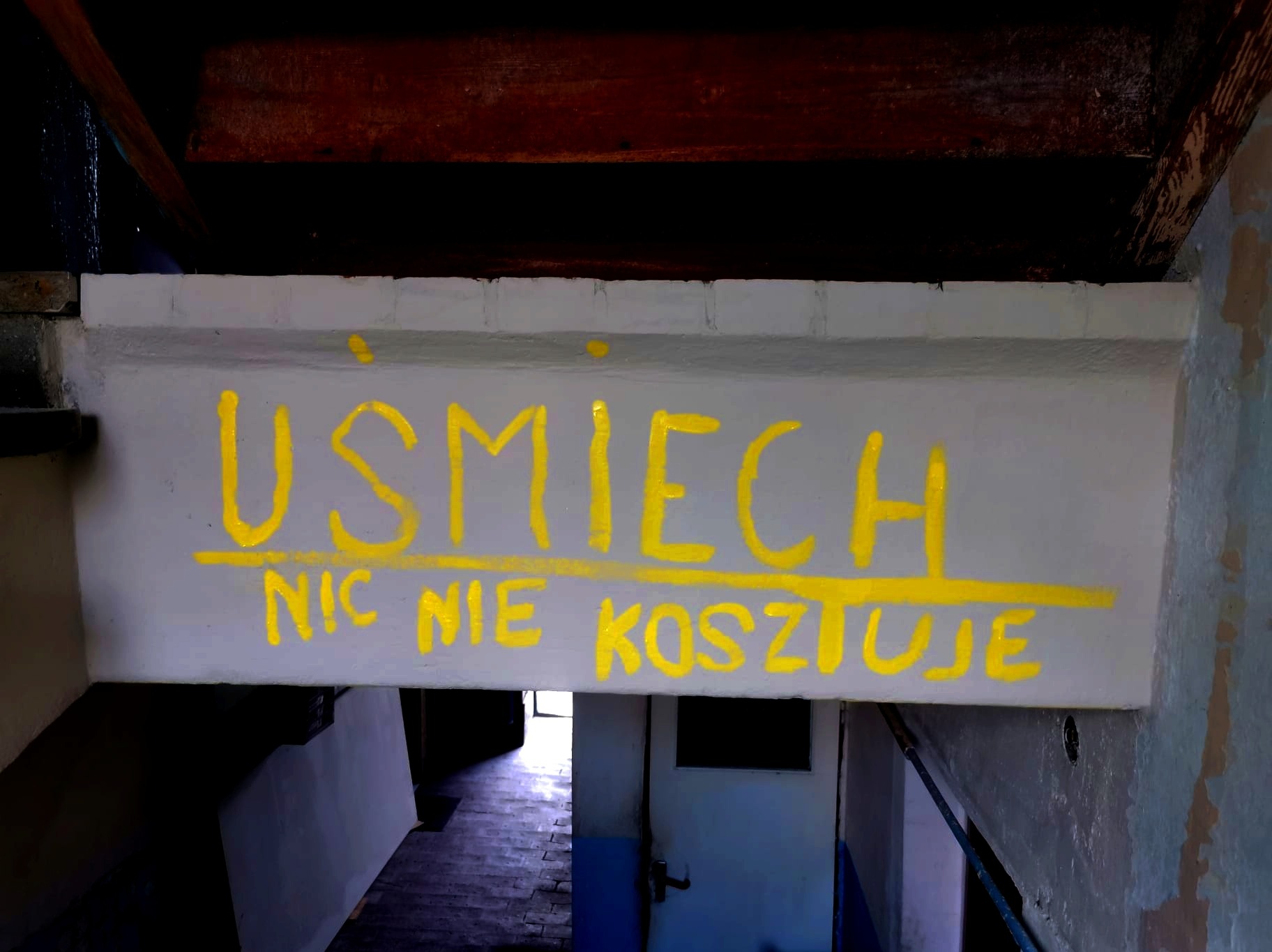 Żółty napis 'Uśmiech nic nie kosztuje' namalowany na szarym betonowym nadprożu w klatce schodowej. Widoczne drewniane belki stropowe i schody prowadzące w dół.