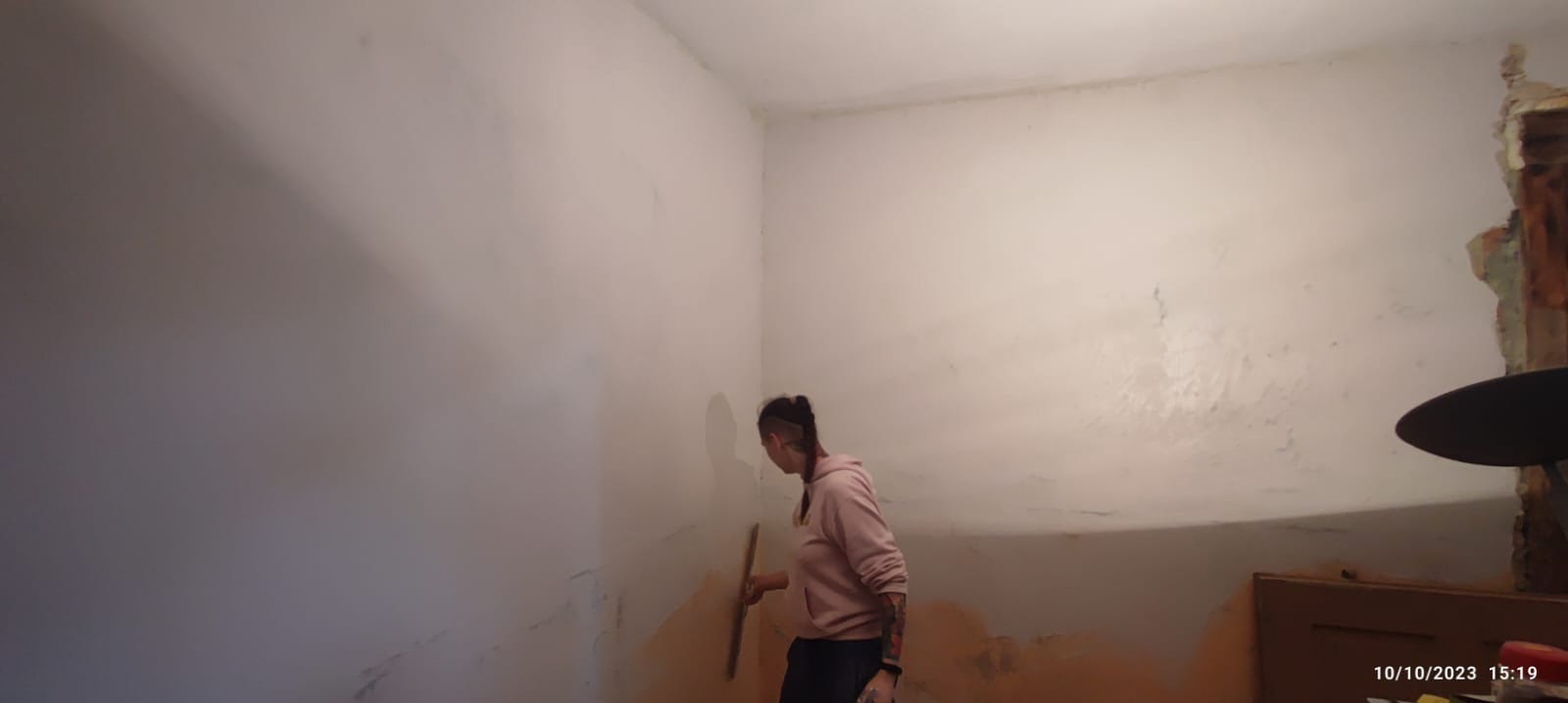 Kobieta z tatuażami na ramieniu wyrównuje ścianę szpachelką w pomieszczeniu w trakcie remontu, widoczny fragment zniszczonej ściany z cegłami.