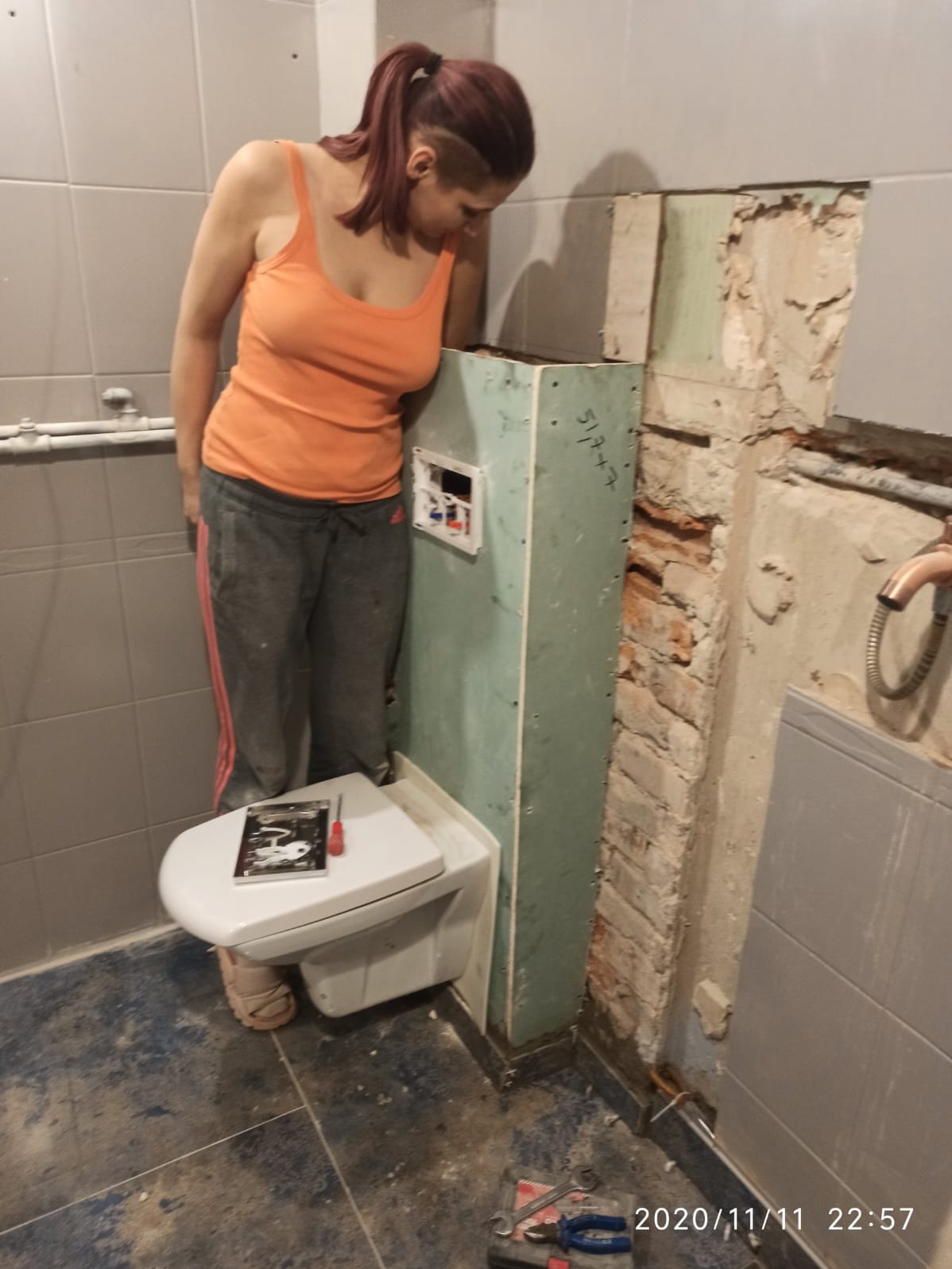 Kobieta w pomarańczowym podkoszulku i dresach montuje podtynkowy stelaż WC w trakcie remontu łazienki, widoczne odsłonięte cegły i narzędzia.
