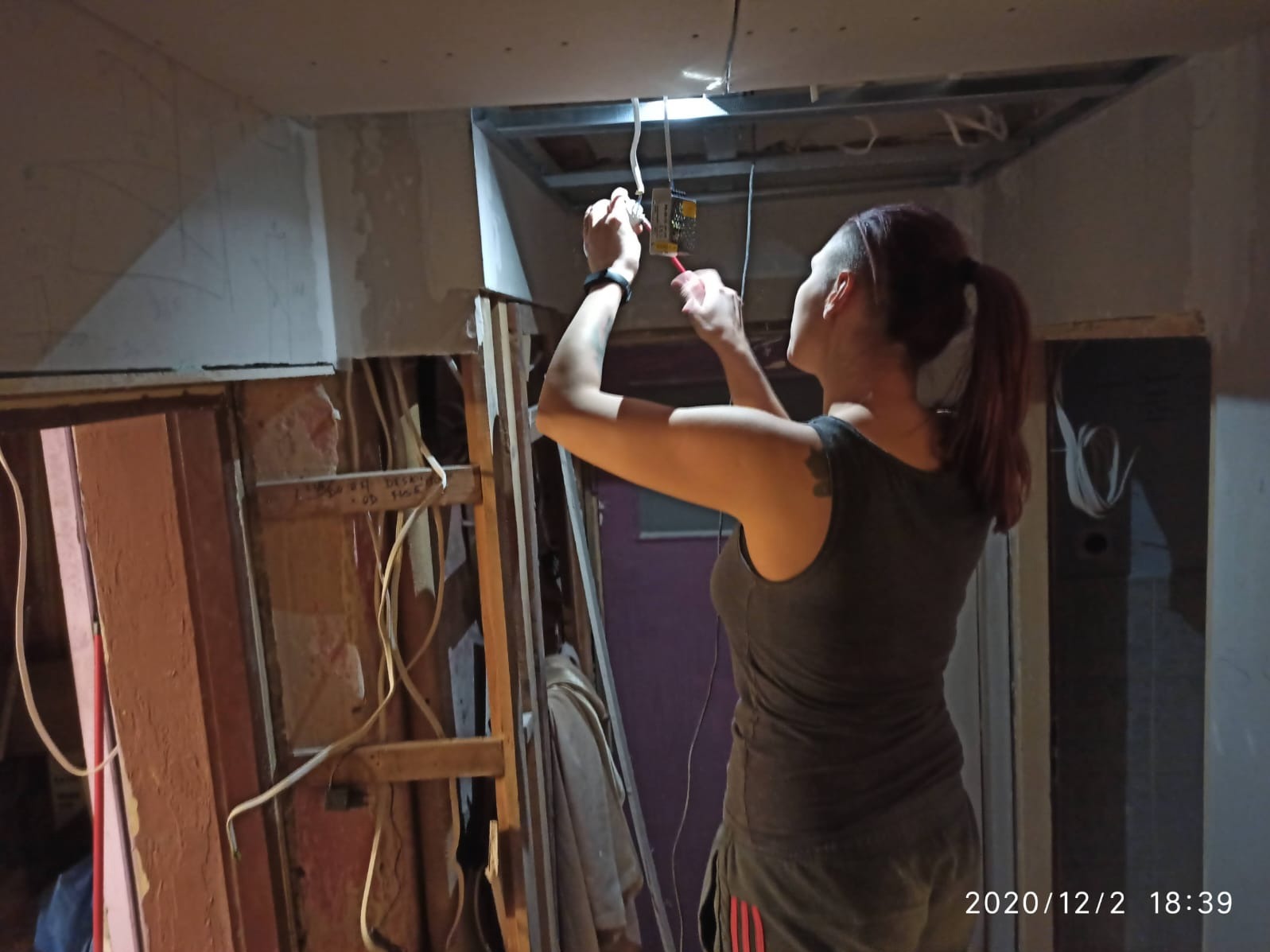 Kobieta z włosami spiętymi w kucyk, w szarym podkoszulku i krótkich spodenkach, pracuje przy instalacji elektrycznej pod sufitem w trakcie remontu pomieszczenia. Widoczne odsłonięte przewody...