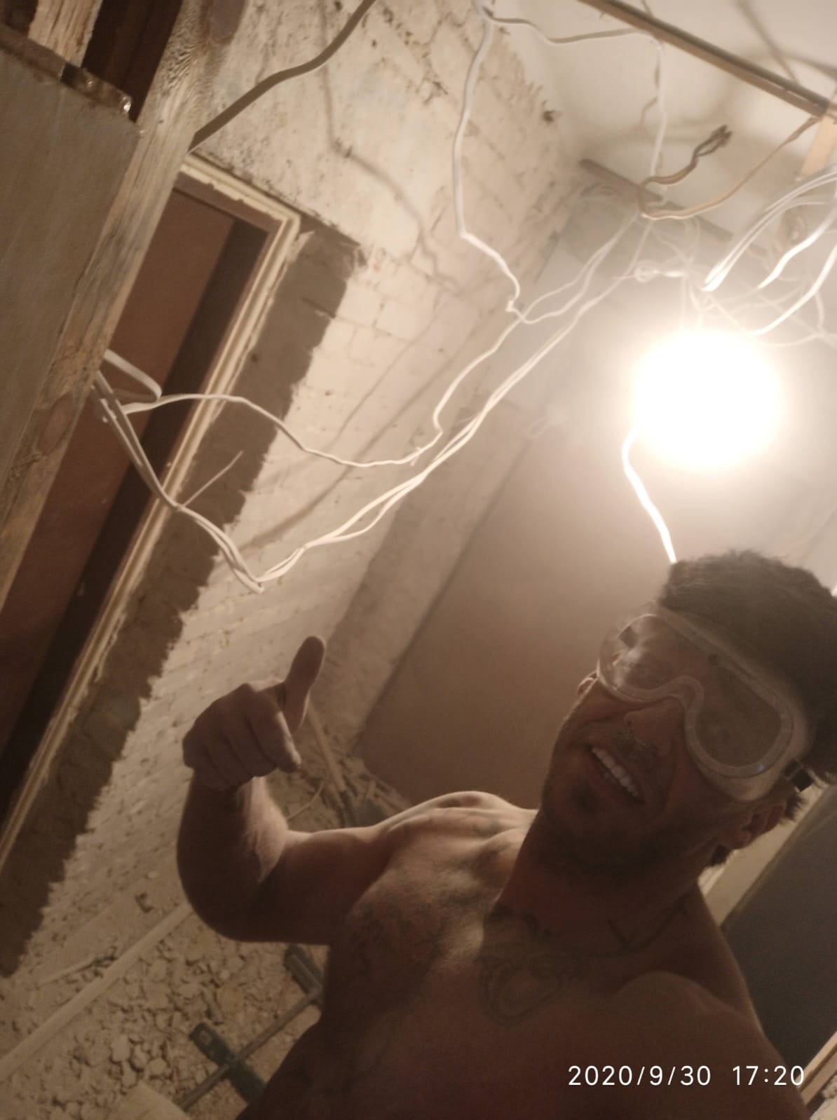 Uśmiechnięty mężczyzna bez koszulki w okularach ochronnych, pokazujący kciuk w górę w trakcie prac instalacyjnych w pomieszczeniu z odsłoniętą cegłą i widocznymi przewodami elektrycznymi pod sufitem.