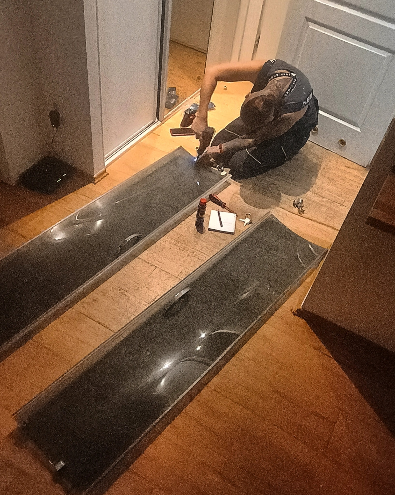 Monterażysta w trakcie pracy nad szklanym panelem, używając wkrętarki, na podłodze leżą dwa panele, notatnik, klucze i narzędzia.