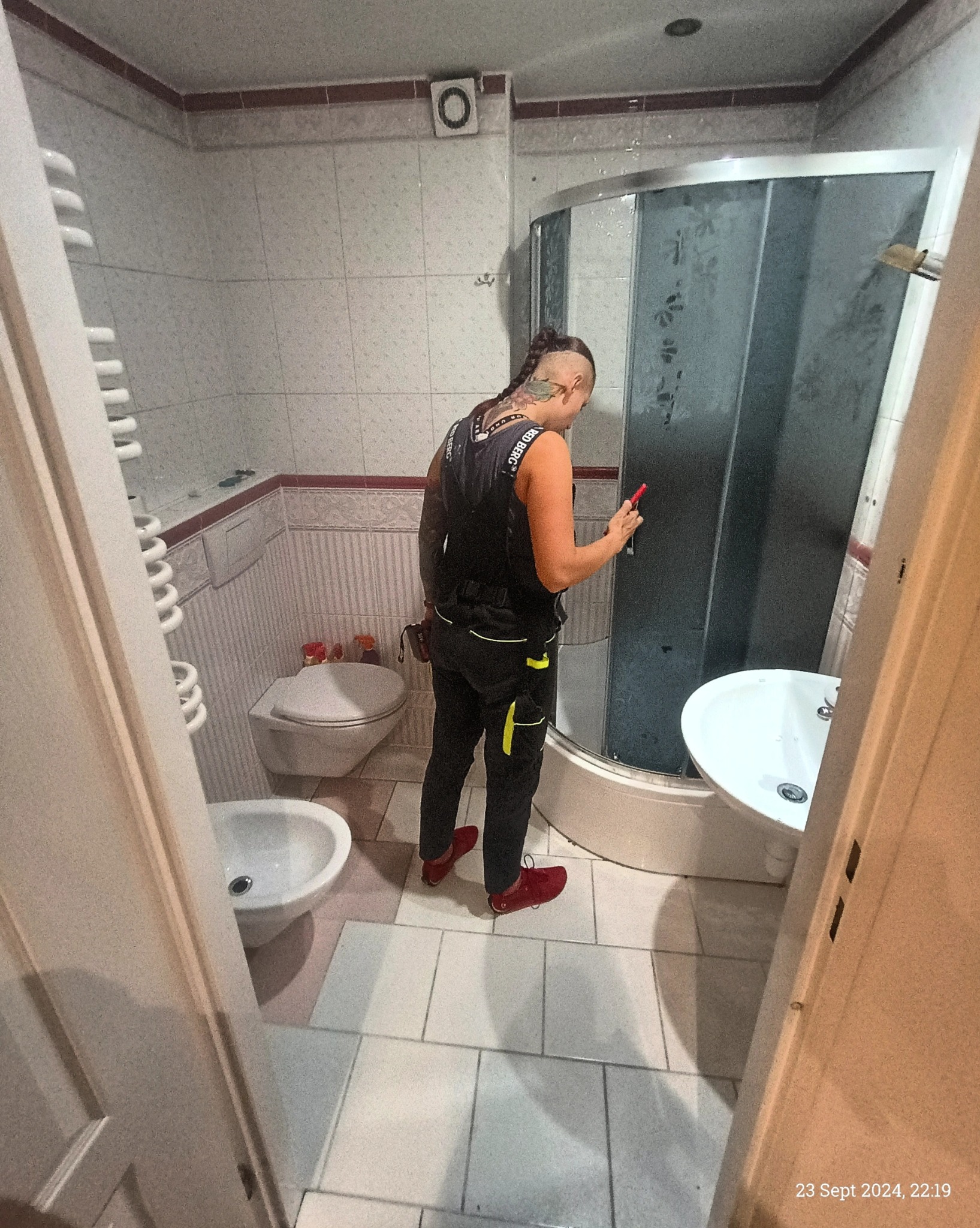 Osoba w roboczym stroju, z częściowo ogoloną głową i tatuażami, stoi w łazience z telefonem w ręku, prawdopodobnie sprawdzając postęp prac przy kabinie prysznicowej, widoczne elementy wyposażenia...