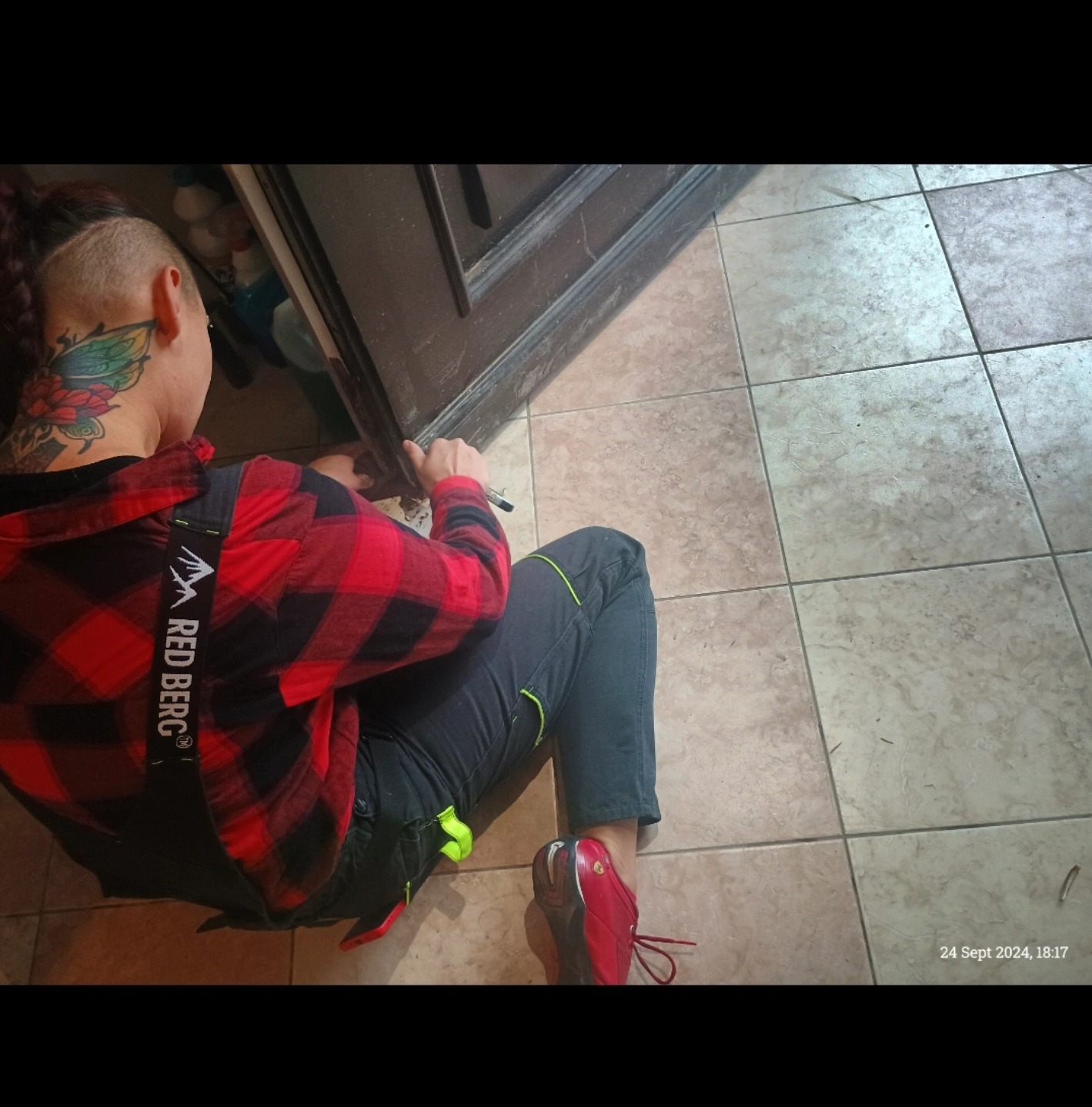 Osoba w czerwonej koszuli w kratę, siedząca na podłodze z płytek i dopasowująca dolną krawędź ciemnych drzwi, z widocznym tatuażem na szyi.