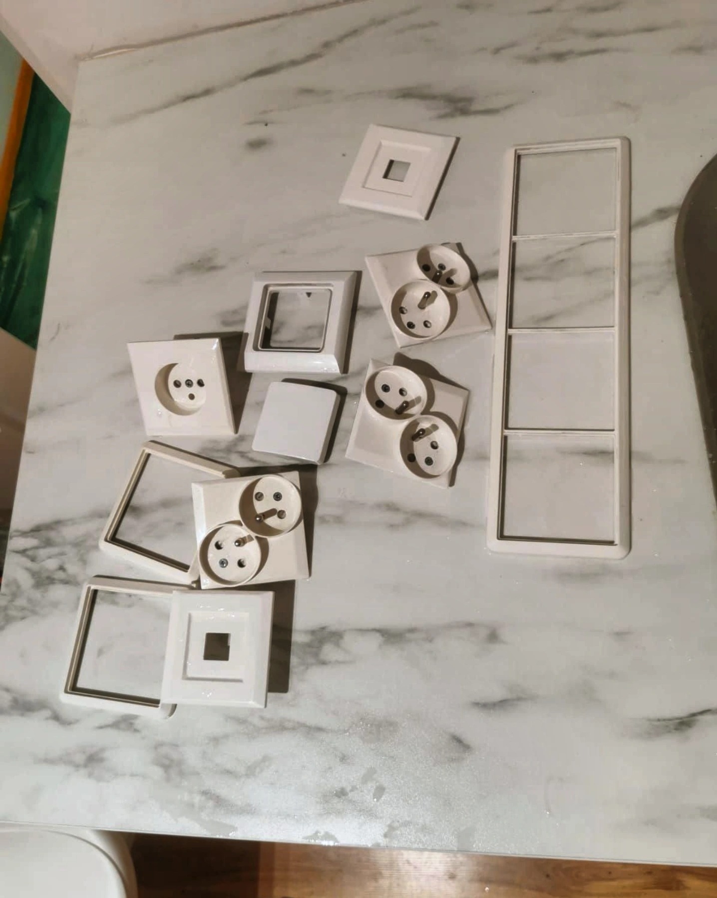 Rozłożone, białe elementy gniazdek i włączników elektrycznych, w tym ramki pojedyncze i potrójne, leżące na blacie o marmurkowej fakturze.