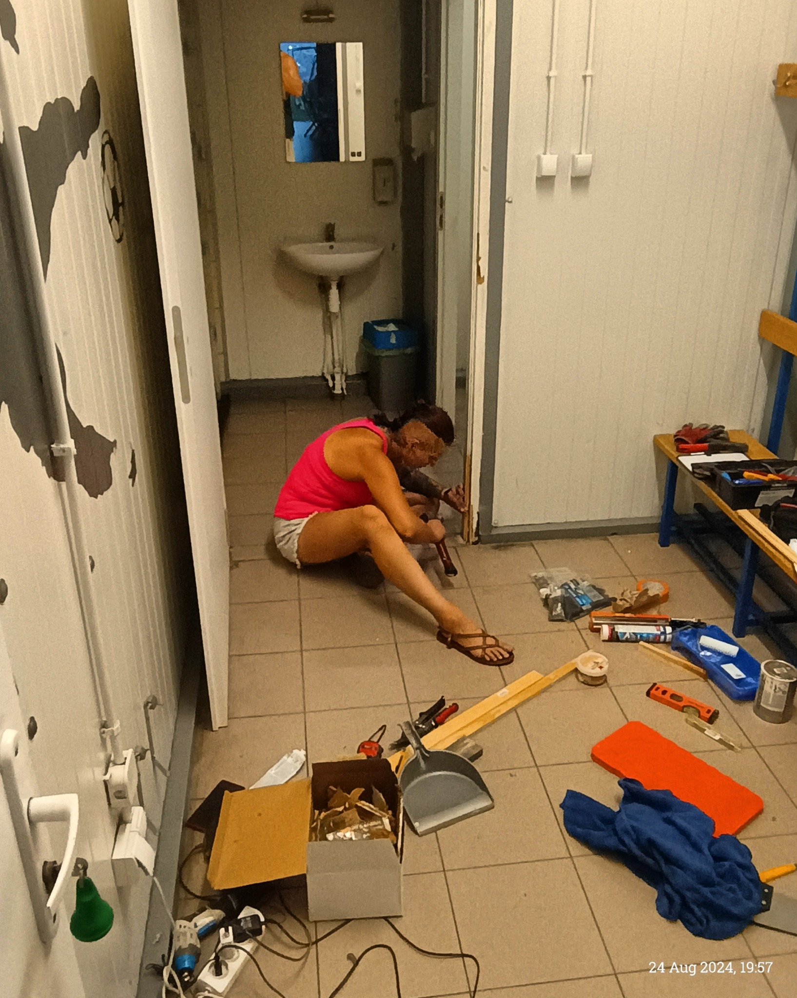 Kobieta w różowej koszulce i sandałach siedzi na podłodze w pomieszczeniu, prawdopodobnie podczas prac remontowych, używając narzędzia do obróbki drewna przy drzwiach. Wokół niej rozrzucone...