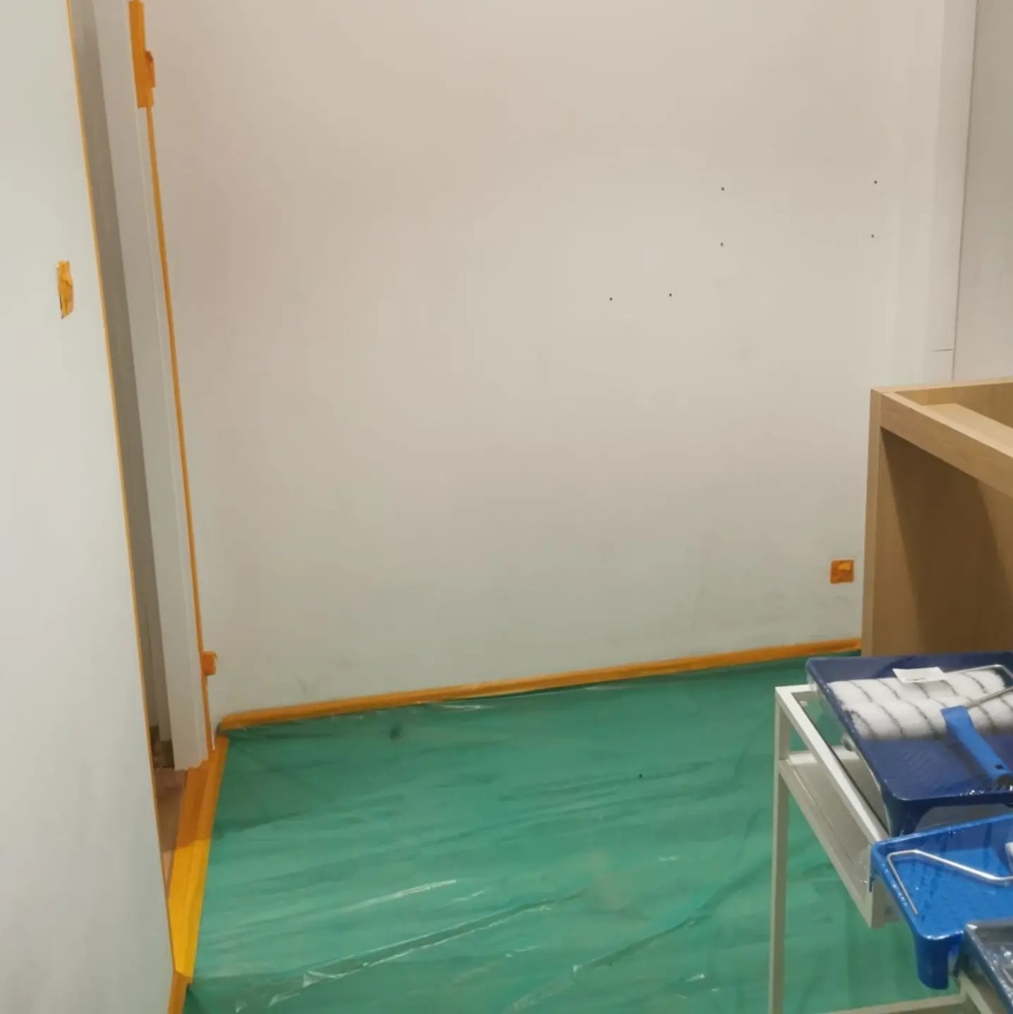 Przygotowanie do malowania ścian: zabezpieczona podłoga folią ochronną, taśma malarska na listwach i framugach, wałki malarskie w kuwetach na metalowym stojaku, ściana z widocznymi śladami po...