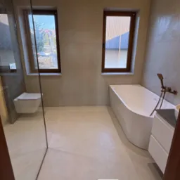 Nowoczesna łazienka z beżowymi płytkami, kabiną prysznicową ze szklaną ścianką, podwieszaną toaletą, białą wanną i oknami z drewnianymi ramami.