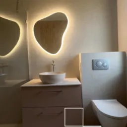 Nowoczesna łazienka z podświetlanym lustrem o nieregularnym kształcie, umywalką nablatową i podwieszaną toaletą. Beżowe płytki na ścianach i podłodze.