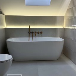 SG Modern Grzegorz Stopka - Minimalistyczna łazienka na poddaszu z wolnostojącą wanną, złotą armaturą i oknem dachowym. Ściana wyłożona pionowymi, wąskimi płytkami. Subtelne oświetlenie LED.