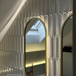 SG Modern Grzegorz Stopka - Łazienka na poddaszu z owalnymi, szarymi płytkami, złotymi ramami luster i oświetleniem LED. Widoczne okno dachowe i fragment instalacji. Nowoczesny design.
