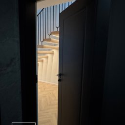 SG Modern Grzegorz Stopka - Ciemne drzwi uchylone, ukazujące schody z podświetleniem LED i parkiet w jodełkę. Minimalistyczny design wnętrza, widok z korytarza.