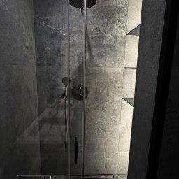 SG Modern Grzegorz Stopka - Nowoczesna kabina prysznicowa z czarnymi profilami i deszczownicą, wykończona szarą płytką imitującą kamień. Widoczne półki wnękowe z podświetleniem.