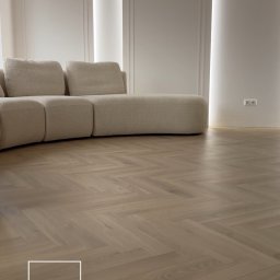 SG Modern Grzegorz Stopka - Jasny parkiet ułożony we wzór jodełki francuskiej, z widoczną sofą w tle i oświetleniem LED w ścianie. Minimalistyczne wnętrze z dbałością o detale.