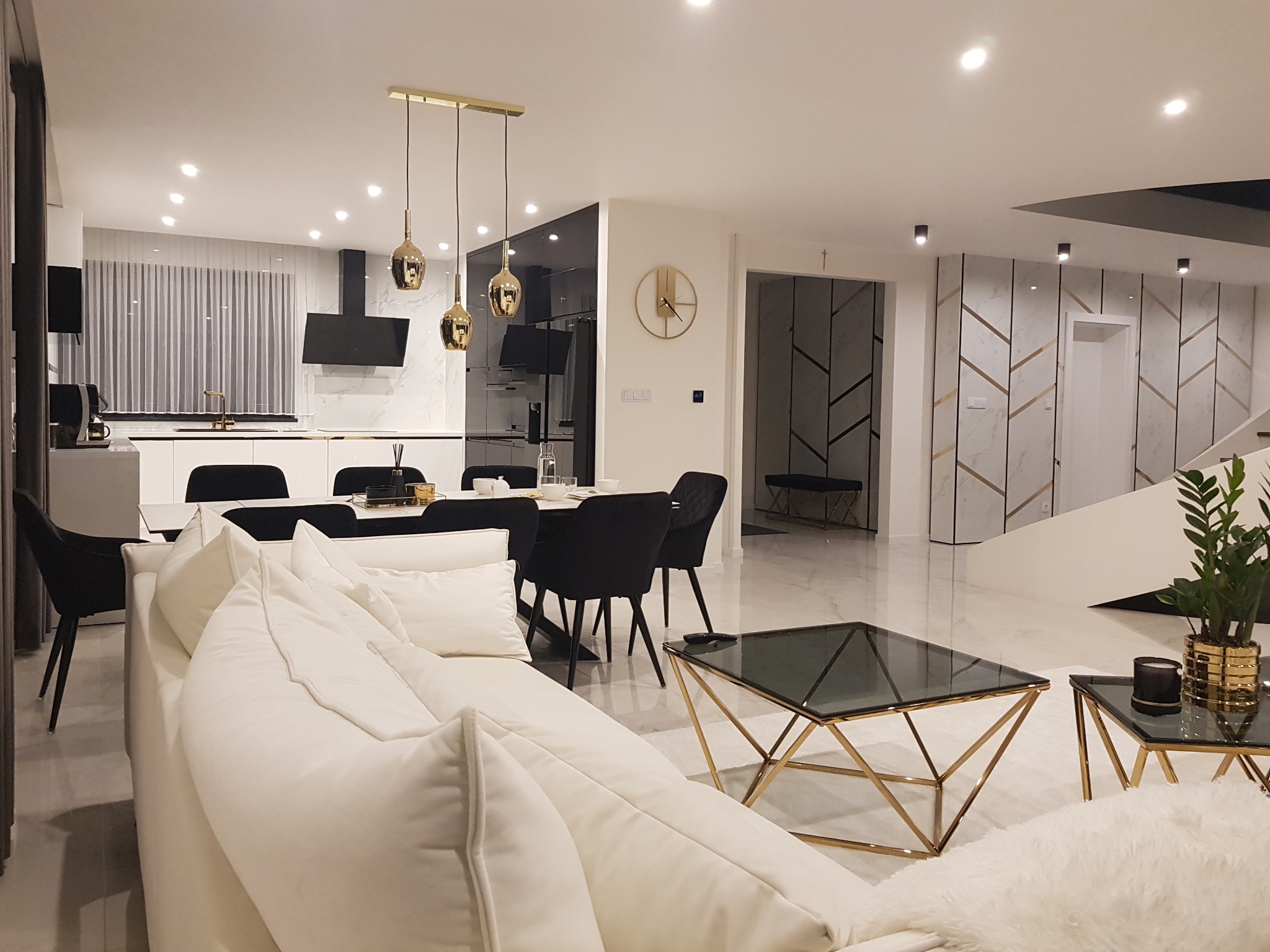 Przestronny salon z białym narożnikiem, szklanymi stolikami kawowymi ze złotymi nogami, jadalnią z czarnymi krzesłami i otwartą kuchnią w tle, całość utrzymana w nowoczesnym stylu.