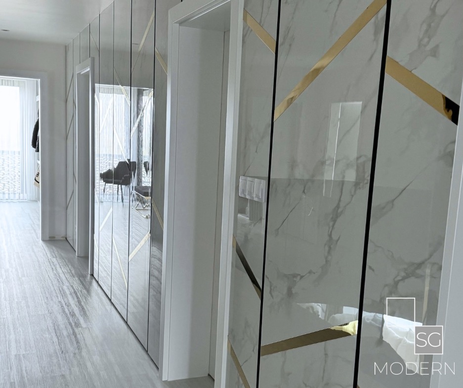 Nowoczesny korytarz z panelami ściennymi imitującymi marmur, zdobionymi złotymi paskami, z widocznymi drzwiami i podłogą w odcieniach szarości.