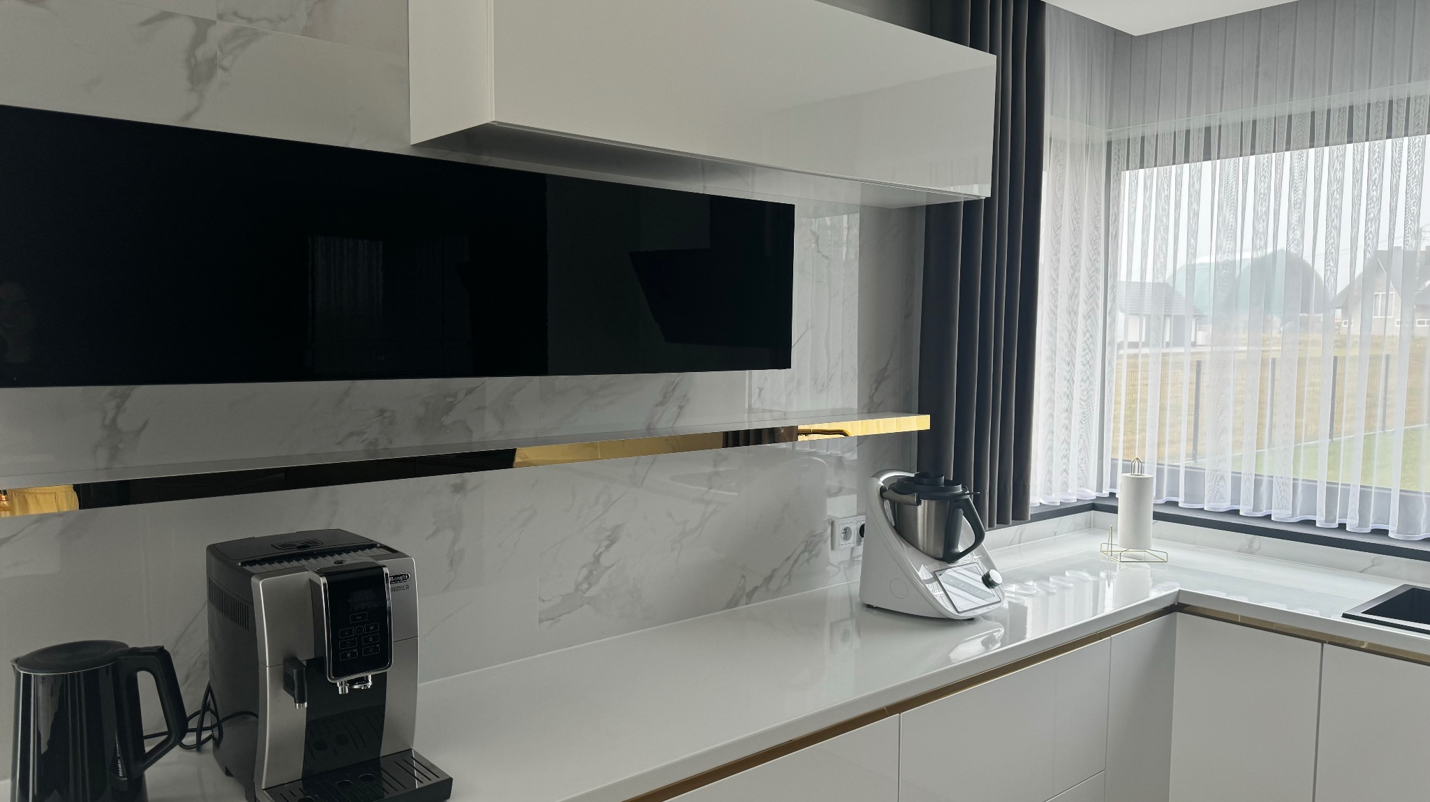 Nowoczesna kuchnia z białymi szafkami z subtelnymi złotymi akcentami, marmurowym backsplashiem, czarnym panelem nad blatem, oraz sprzętami AGD: ekspres do kawy i robot kuchenny. Okno z zasłonami...