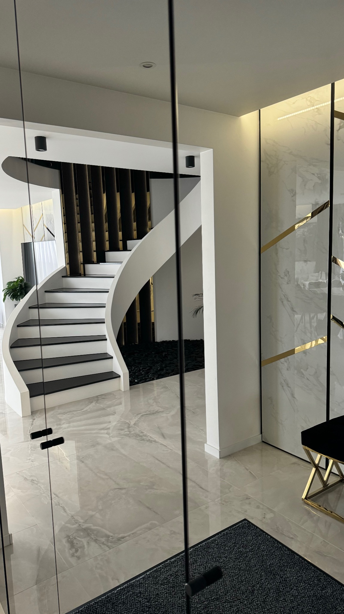 Eleganckie wnętrze z marmurową podłogą, spiralnymi schodami z czarnymi stopniami i dekoracyjną ścianą z marmuru i złotych akcentów, widok przez szklane drzwi.