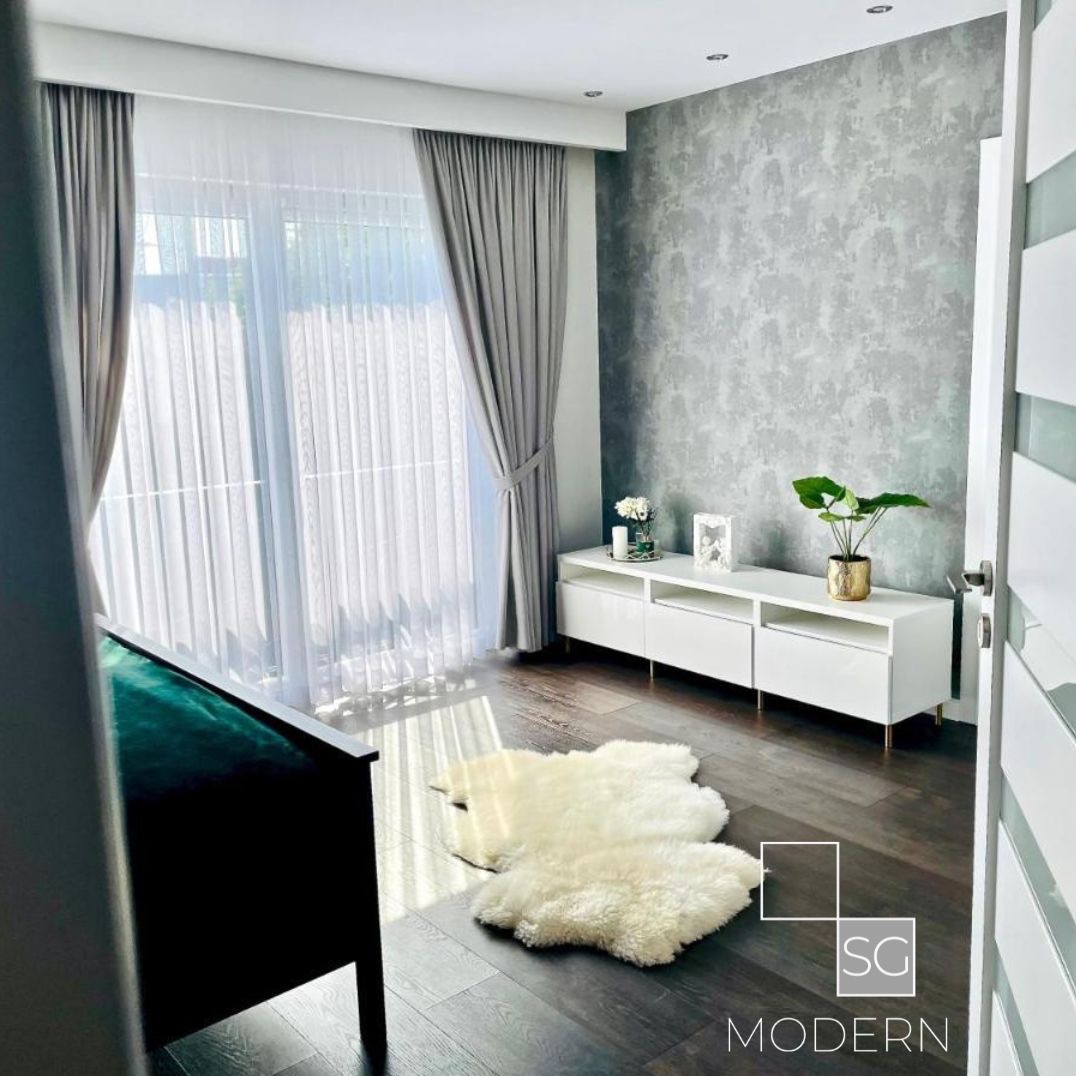 Nowoczesny salon z szarą, teksturowaną ścianą, białymi meblami, puszystym dywanem i zasłonami w odcieniach szarości, doświetlony naturalnym światłem.