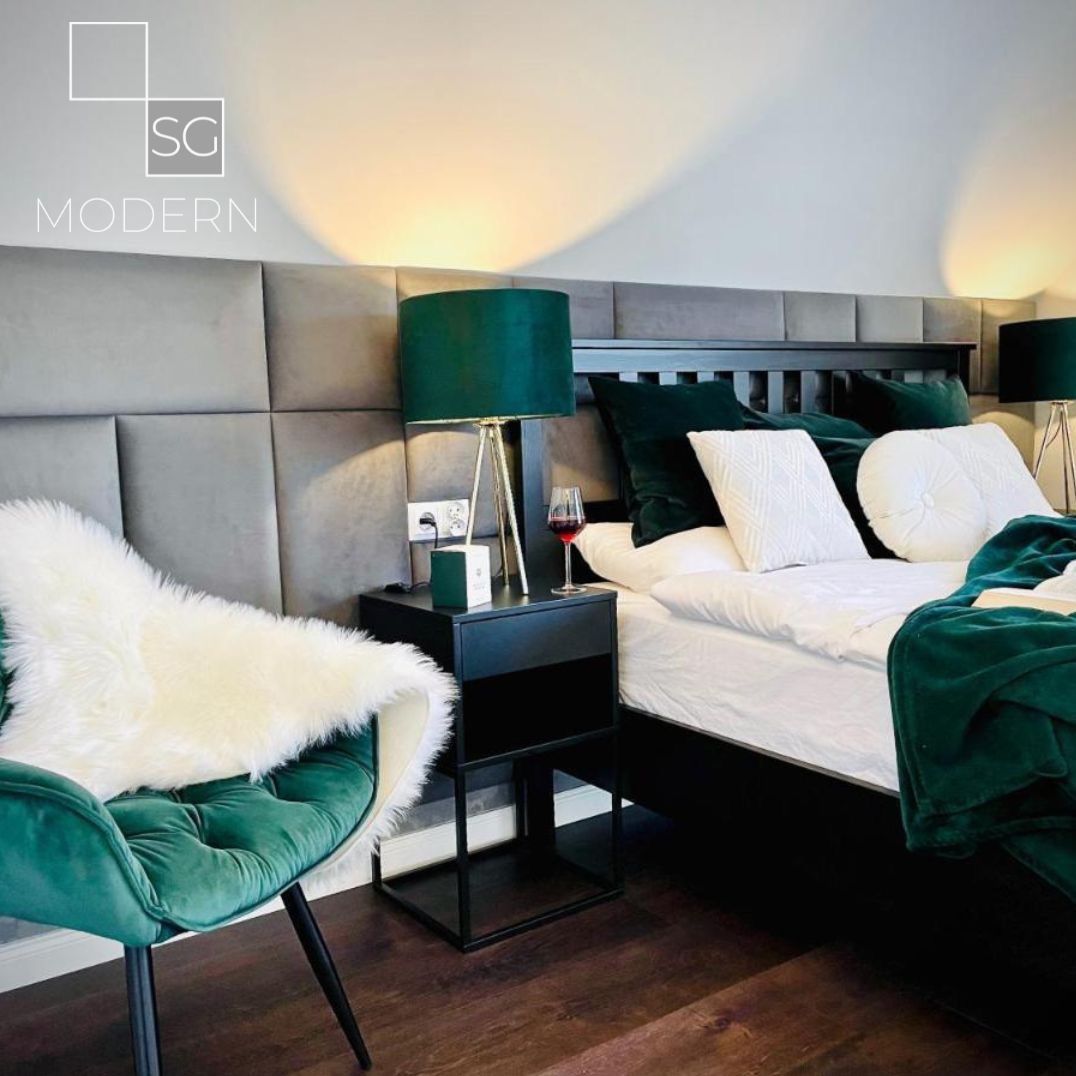Nowoczesna sypialnia z szarą tapicerowaną ścianą, ciemnozielonymi akcentami i stolikiem nocnym z lampką, uzupełniona kieliszkiem czerwonego wina.