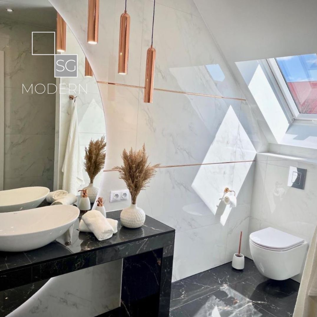 Nowoczesna łazienka na poddaszu z marmurowymi płytkami, czarnym blatem, owalną umywalką, miedzianymi lampami wiszącymi i oknem dachowym wpuszczającym jasne światło.