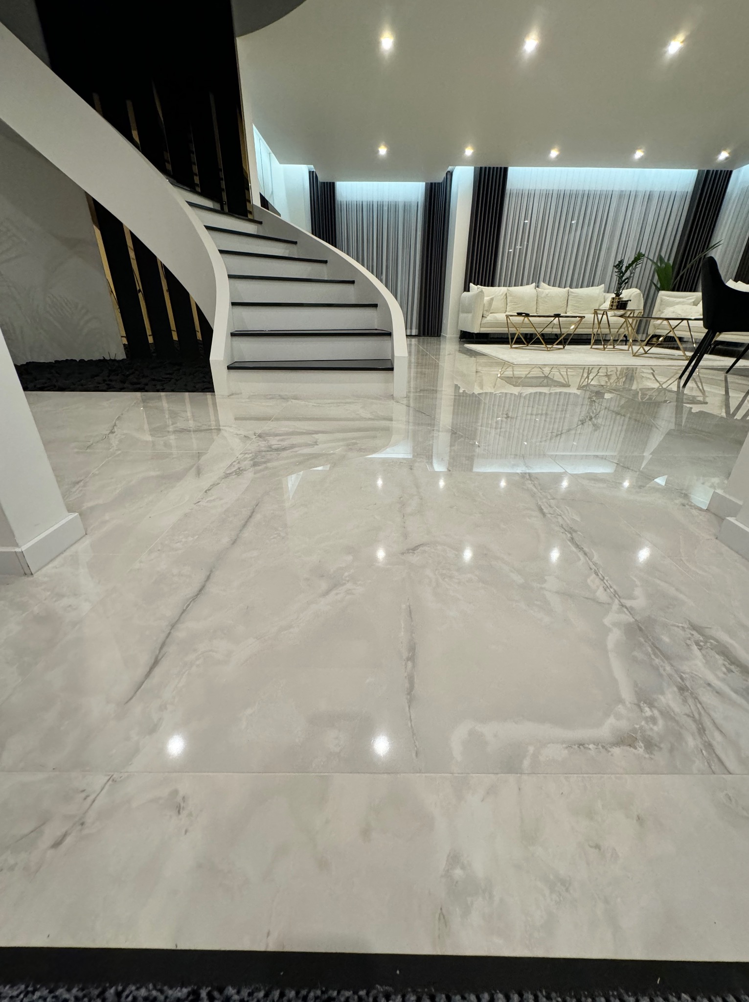 Błyszcząca podłoga z jasnych płytek imitujących marmur, widoczne schody z białymi balustradami i czarnymi akcentami, w tle salon z sofą i stolikami kawowymi.