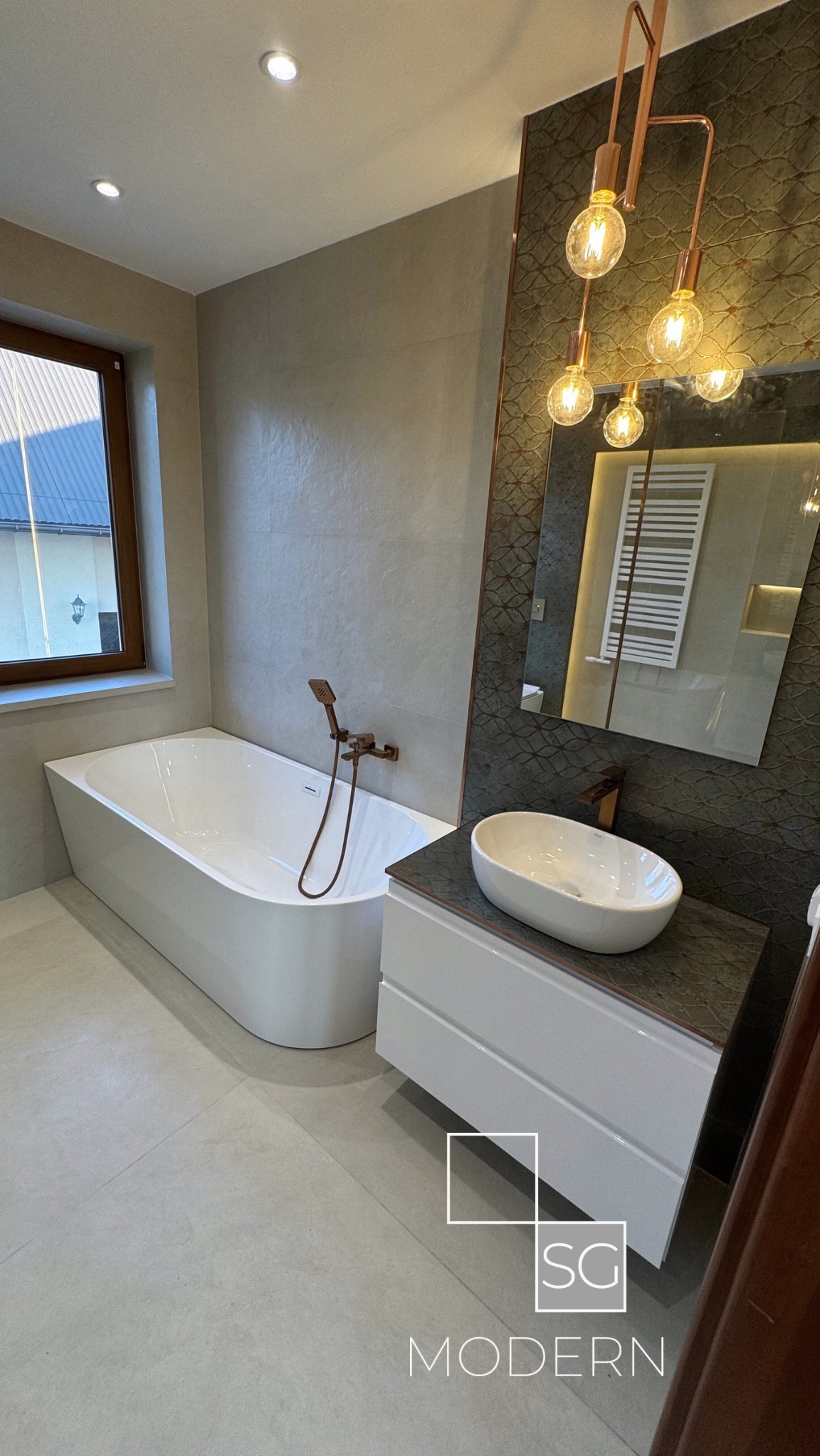 Nowoczesna łazienka z wolnostojącą białą wanną, umywalką nablatową na białej szafce i baterią w kolorze miedzianym, oświetlona wiszącą lampą z żarówkami Edisona i z lustrem w tle z widocznym...