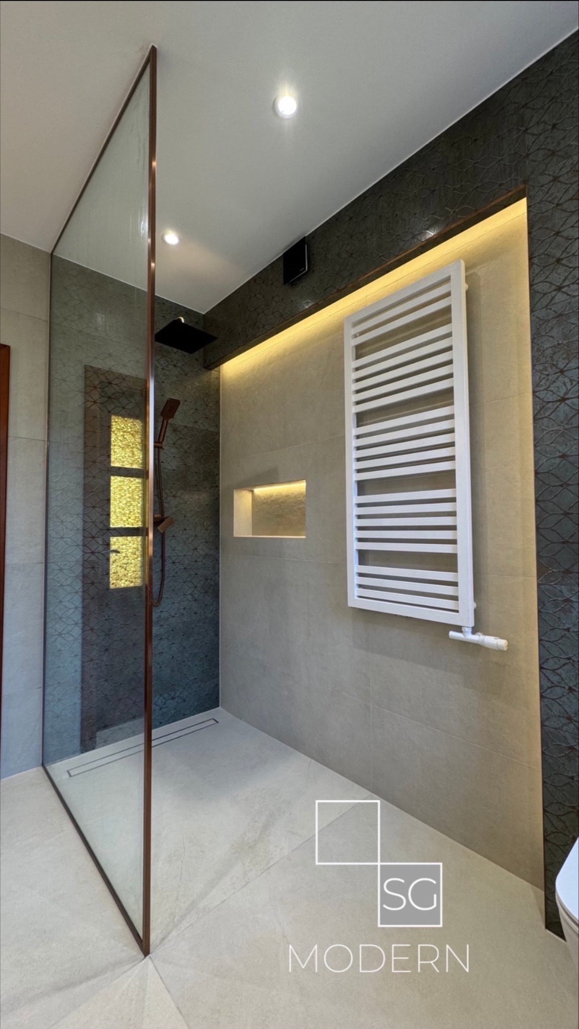 Nowoczesna łazienka z kabiną prysznicową typu walk-in z brązowymi ramami, grzejnikiem drabinkowym, wnęką z podświetleniem oraz płytkami imitującymi wzór liści na jednej ze ścian.