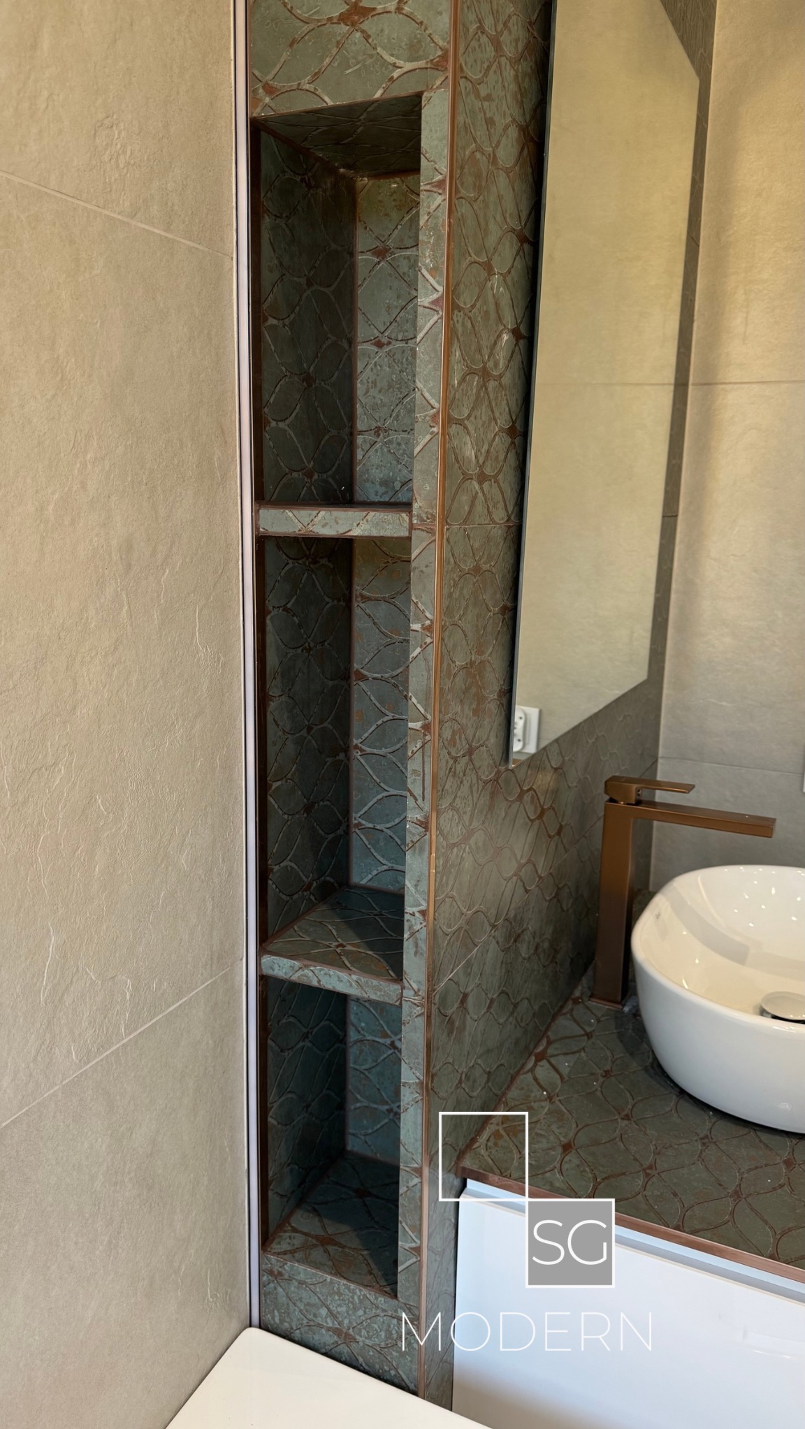 Wykończona łazienka z wnęką z półkami, płytki w odcieniach szarości i zieleni z wzorem, biała umywalka nablatowa i bateria w kolorze miedzi.