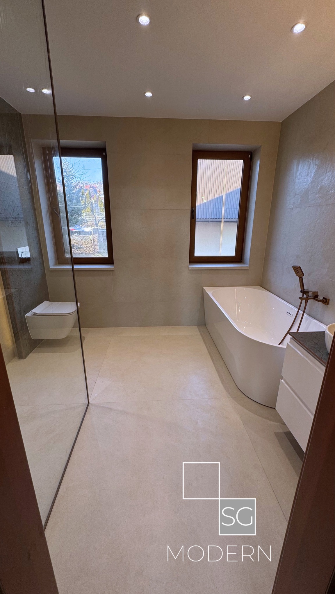 Nowoczesna łazienka z beżowymi płytkami, kabiną prysznicową ze szklaną ścianką, podwieszaną toaletą, białą wanną i oknami z drewnianymi ramami.