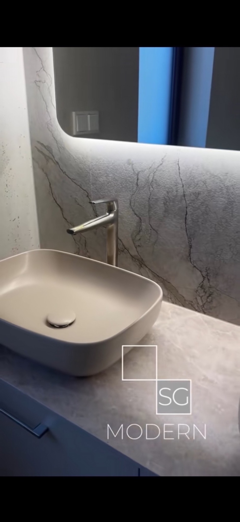 Beżowa umywalka nablatowa z chromowaną baterią na blacie z efektem marmuru. W tle lustro z podświetleniem LED i marmurowe wykończenie ściany. Nowoczesny design łazienki.