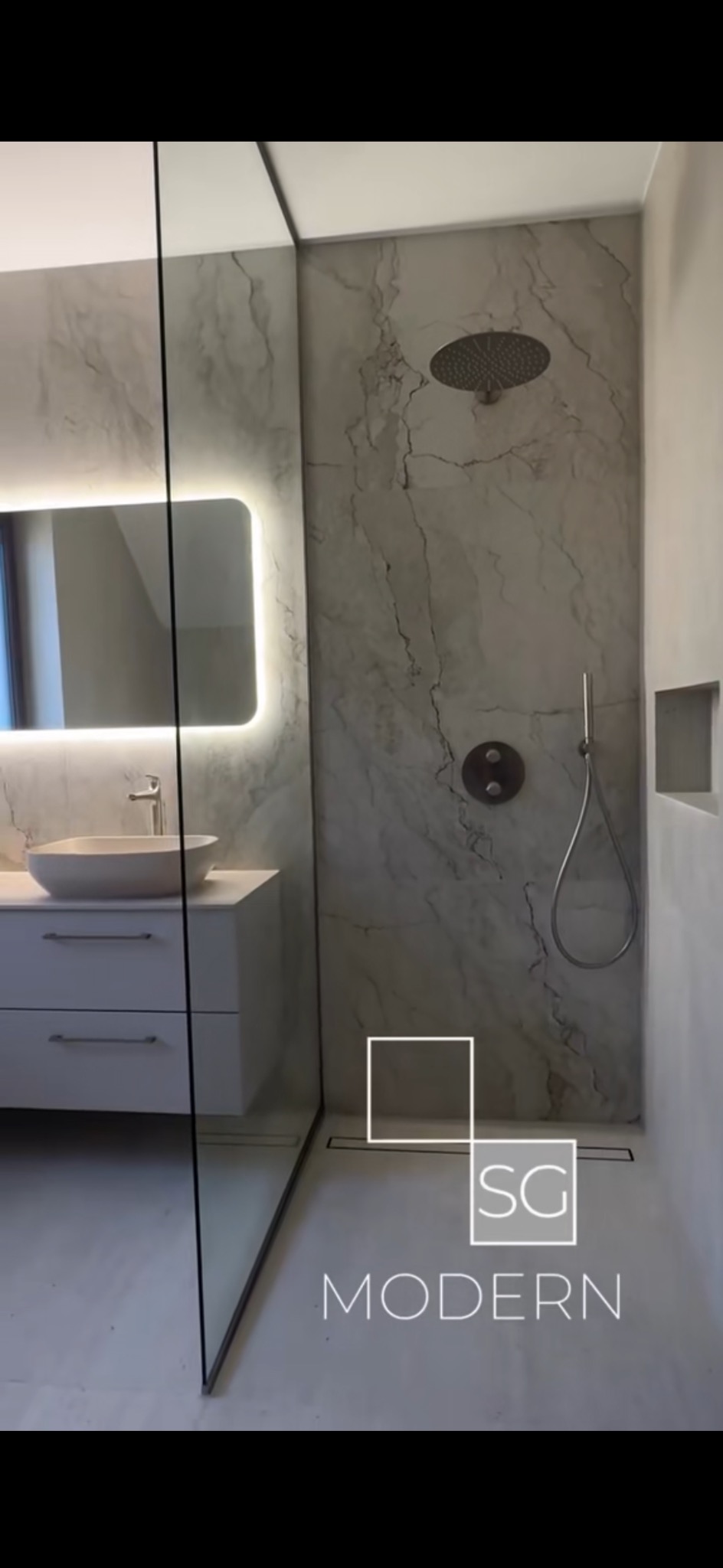 Nowoczesna łazienka z marmurowymi płytkami, deszczownicą i podświetlanym lustrem nad umywalką. Minimalistyczny design z kabiną prysznicową typu walk-in.