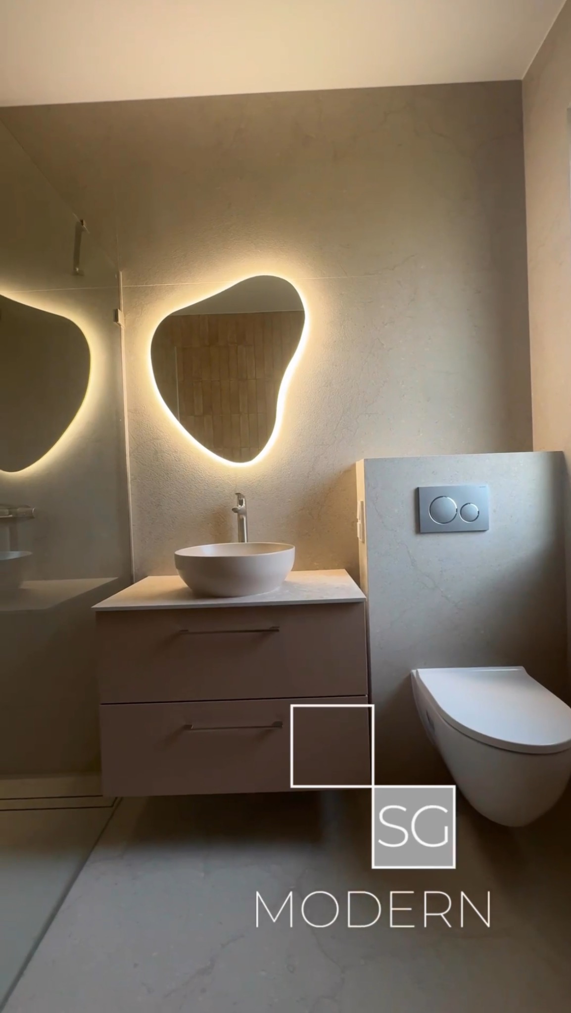 Nowoczesna łazienka z podświetlanym lustrem o nieregularnym kształcie, umywalką nablatową i podwieszaną toaletą. Beżowe płytki na ścianach i podłodze.